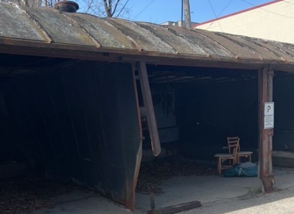 Deteriorating open-air carport structure with rusted roof and leaning wooden supports.